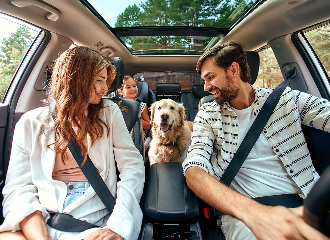 Contact - Smiling Young Parents Looking Back at Their Dog in the Back Seat While Driving a Car During a Road Trip with Their Daughter on a Sunny Day